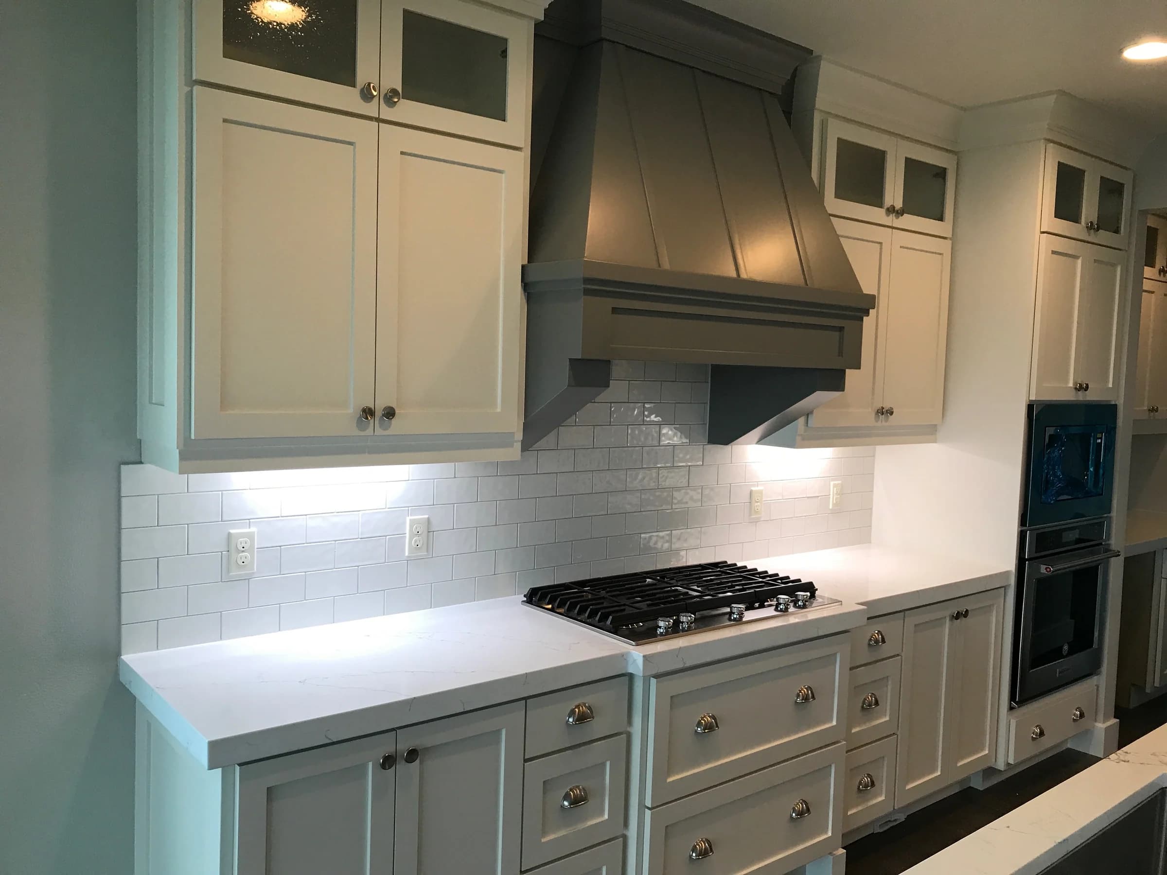 NS Wood Finishing project: light painted cabinetry with glass-front upper doors beside a gray range hood
